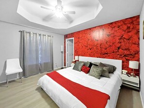A bedroom with a bed, a ceiling fan, a red floral wall, a white chair, and a door.