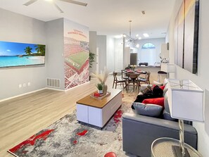 A modern living room with a large TV, a dining area, and a kitchen in the background.