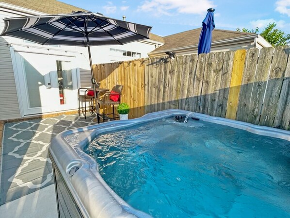 A hot tub in a backyard with a wooden fence, a patio umbrella, and outdoor furniture.