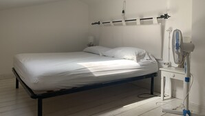 Un dormitorio minimalista con una cama individual, una mesita de noche, un estante en la pared, un ventilador de techo y una ventana que deja entrar luz natural.