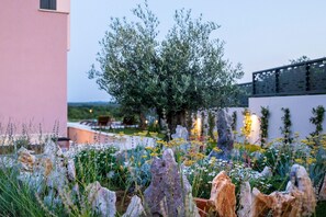 A garden with various plants and rocks, a large tree, and a building in the background.