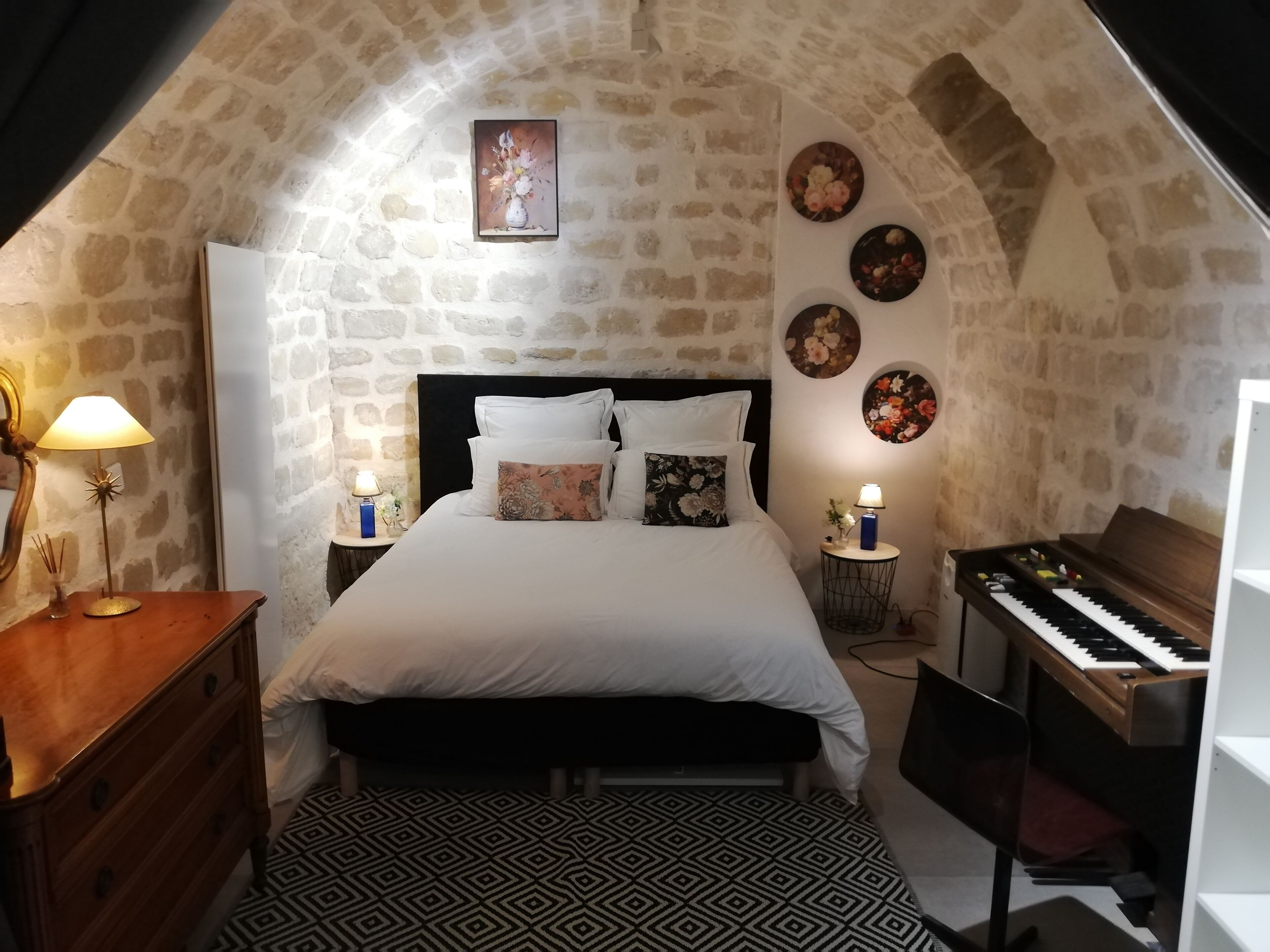 A bedroom with a stone wall, a bed with white bedding, a piano, and decorative wall art.