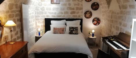 Quarto com parede de pedra, cama com roupa de cama branca, um piano e arte decorativa na parede.