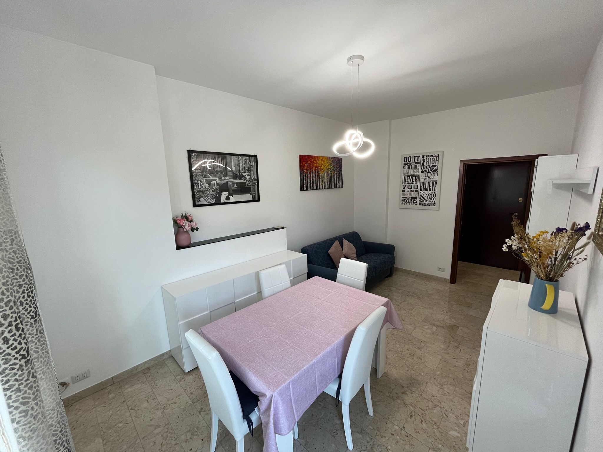 Een moderne woonkamer met een eettafel, witte stoelen, een bank en een lange bank. Achtergrond met kunstwerken en een deur.