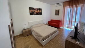 Un dormitori amb un llit, un sofà vermell, un escriptori amb un ordinador i una finestra amb cortines.