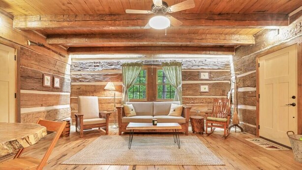 L’intérieur d’un chalet confortable, avec un plancher en bois, un canapé, deux fauteuils, une table à manger et un ventilateur de plafond.