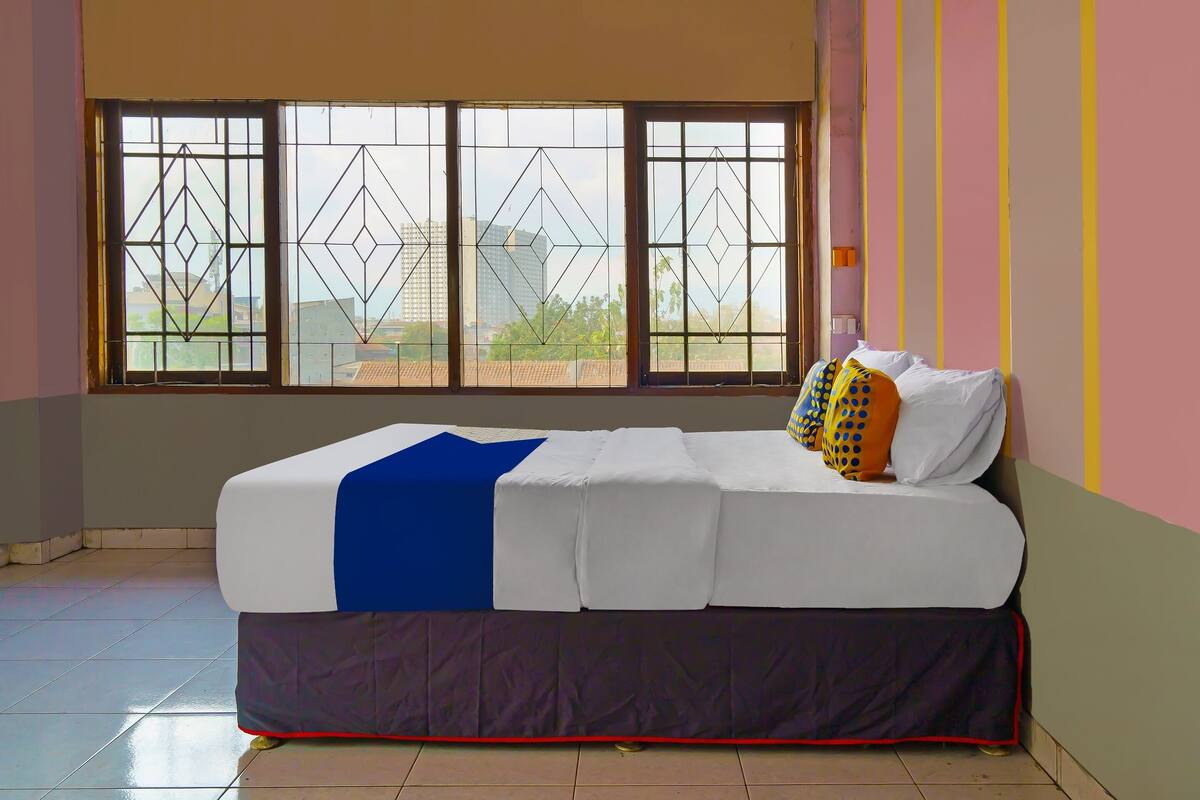 A bed with a blue blanket and white bedding, a window with geometric patterns, and a view of buildings outside.