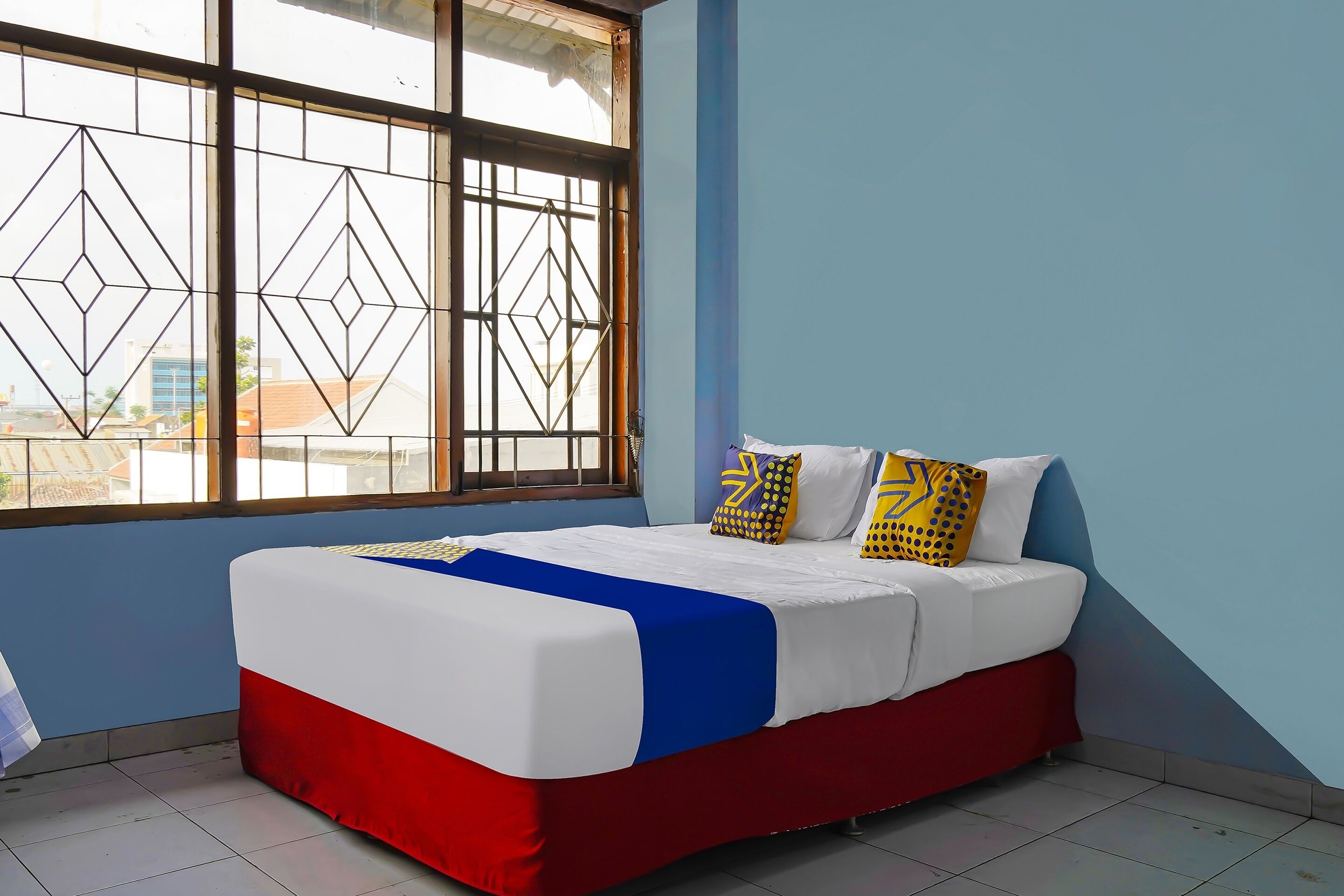 A bedroom with a bed, a window with a geometric pattern, and a view of buildings outside.