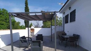 A covered patio area with outdoor furniture, a grill, and a white building with windows and a chimney. - Chalet con Piscina Privada (Chiclana de la Frontera)