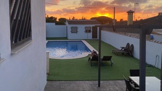 A backyard with a swimming pool, a patio with chairs, and a view of the sunset. - Chalet con Piscina Privada (Chiclana de la Frontera)