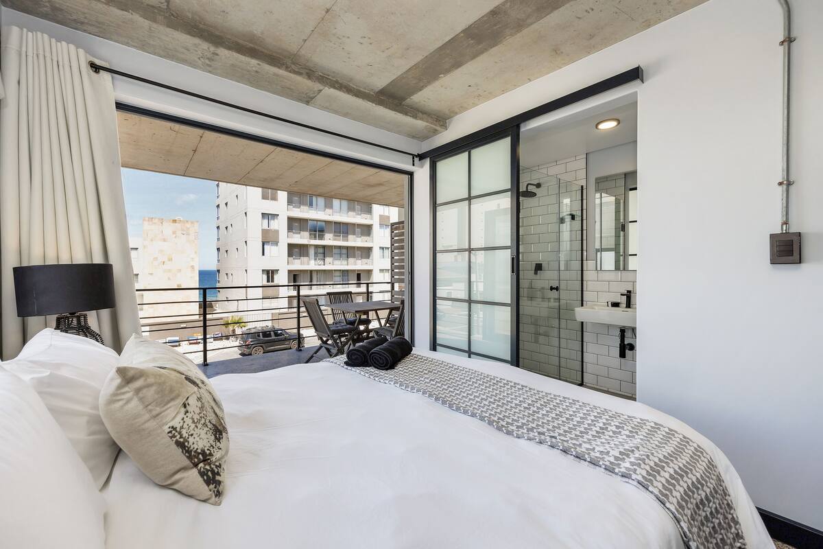 A modern hotel room with a large bed, a view of the city, a balcony with a table and chairs, and a bathroom with a glass shower.