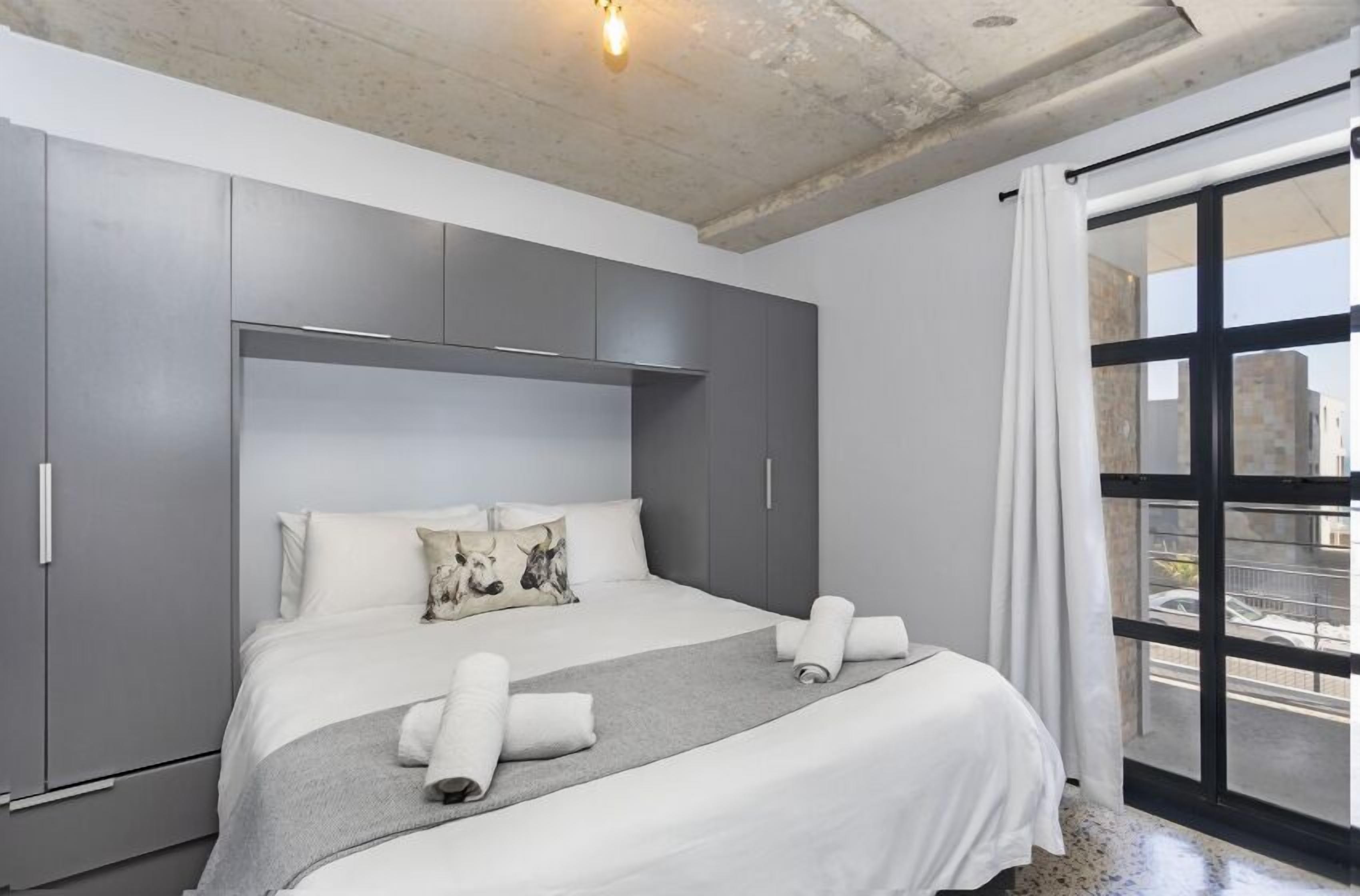 A modern hotel room with a large bed, grey wardrobe, and a view through a large window.