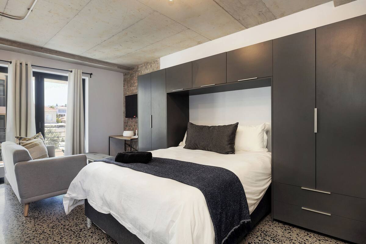A modern bedroom with a large bed, dark wardrobe, and a seating area with a sofa and armchair.
