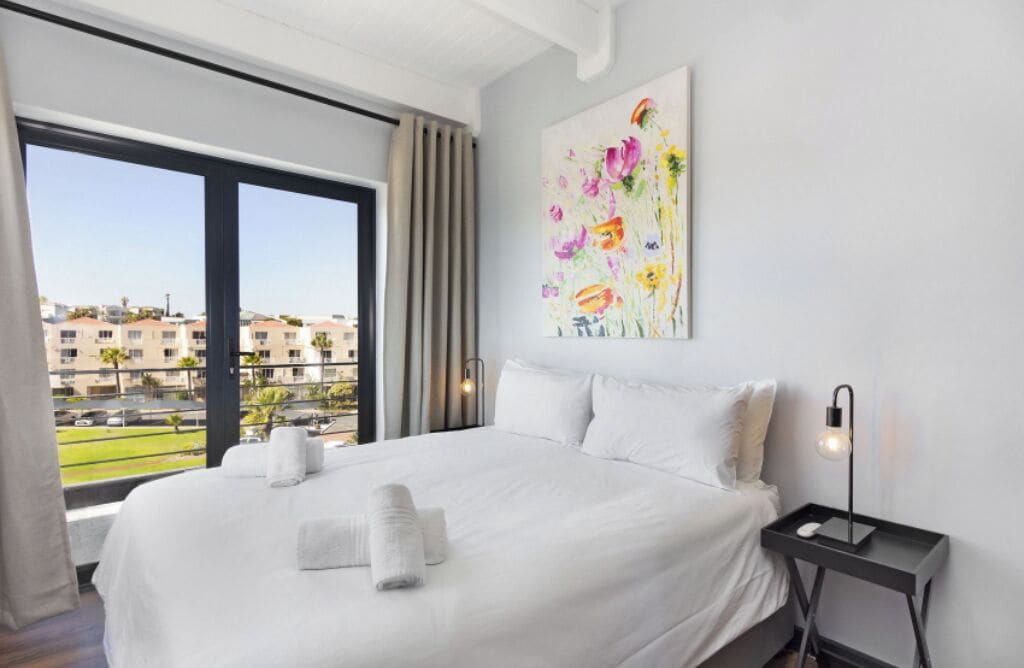 A hotel room with a large bed, a nightstand, a painting, and a view of buildings through the window.