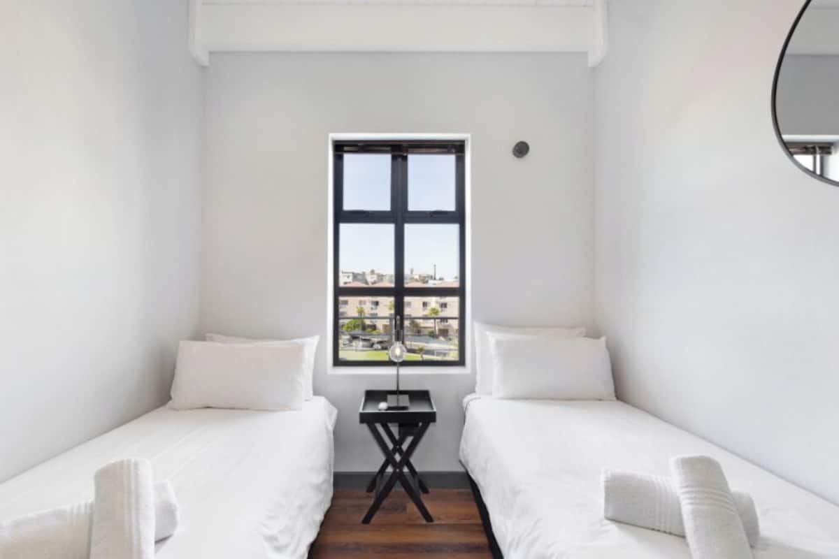 A small, white room with two single beds, a small table, and a window with a view of buildings.