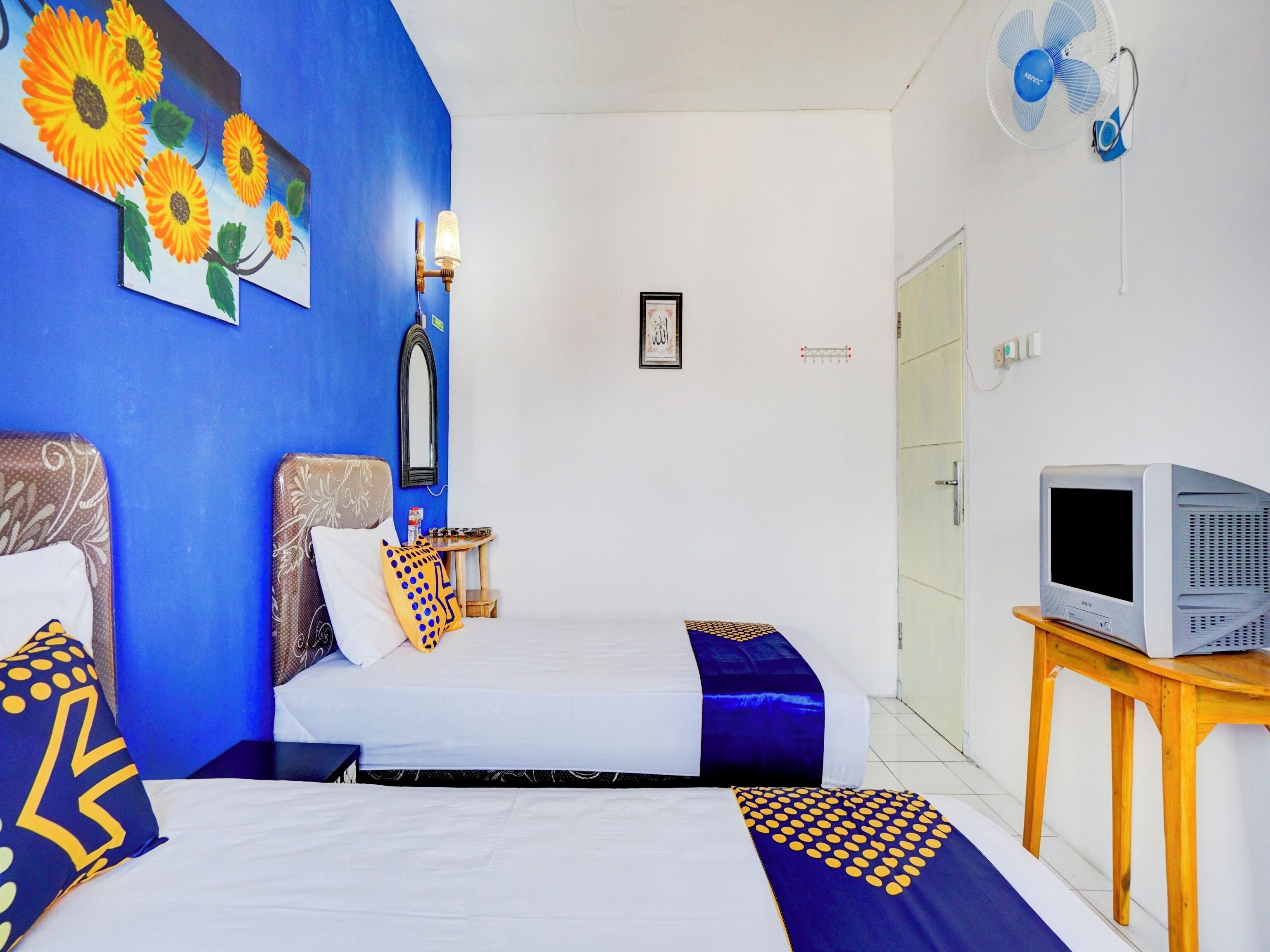 A hotel room with two beds, a TV on a wooden table, a wall-mounted fan, and a painting of sunflowers.