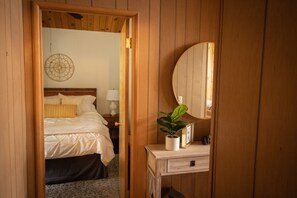 A wooden-paneled room with a bed, a bedside table with a plant, and a mirror. - Cabin - Rustic Glam + Game Room + Views +Workspace- Minutes from attractions (Running Springs)