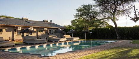 A swimming pool with lounge chairs and umbrellas, surrounded by a brick pathway and a hedge.