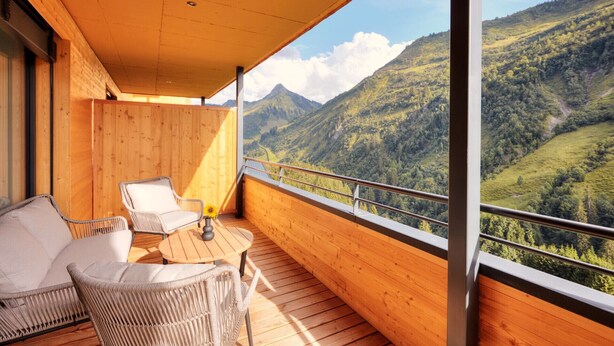 A wooden balcony with wicker furniture and a table, overlooking a mountainous landscape. - Alpenstolz #1.01 Comfort (Damüls)