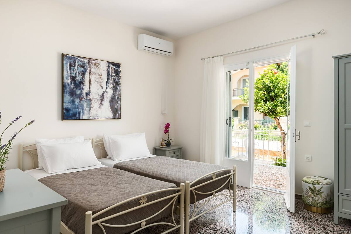 A bedroom with two beds, a small bedside table, a nightstand, an air conditioning unit, a window with curtains, and a view of a courtyard.