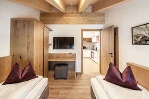 A modern bedroom with wooden beams, a flat-screen TV, a small seating area, and a kitchen in the background. - Alpenstolz #1.09 Exclusive (Damüls)