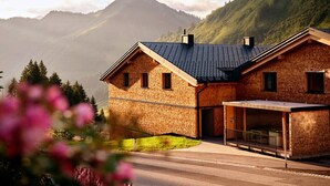 Ein zweistöckiges Ziegelhaus mit einem geneigten Dach, einer Veranda und einem Berg im Hintergrund.