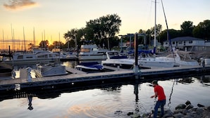 En småbåtshamn med förtöjda båtar, en person som fiskar och en stilla solnedgång i bakgrunden.
