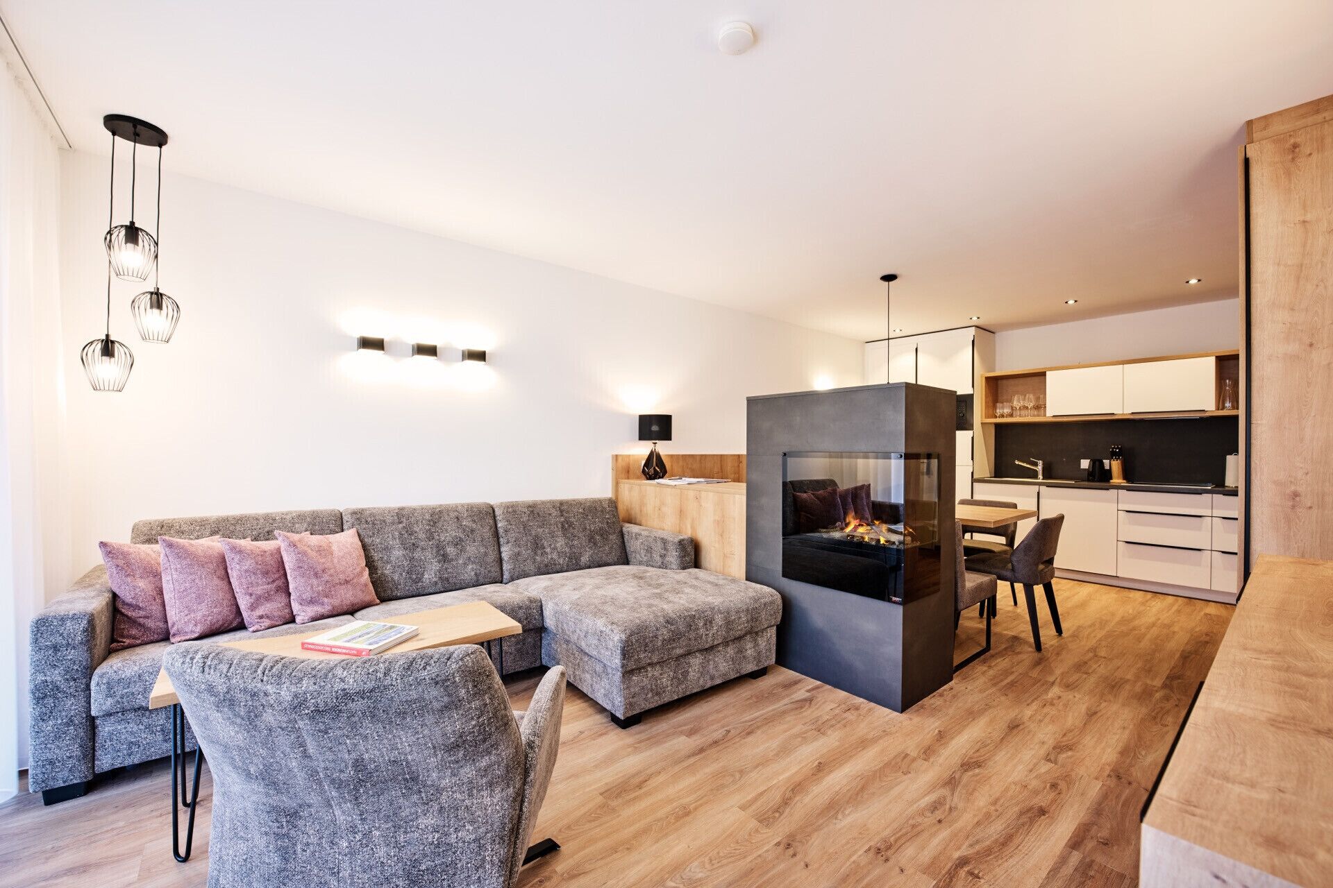 Un salon moderne avec un canapé d’angle gris, un petit pouf et une cheminée. On y trouve également un coin repas avec une table et des chaises, ainsi qu’une cuisine dotée de meubles blancs et de plans de travail en bois.