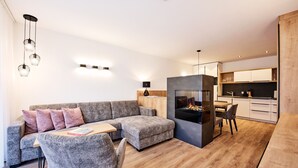 A modern living room with a grey sectional sofa, a small ottoman, and a fireplace. There is a dining area with a table and chairs, and a kitchen with white cabinets and wooden countertops. - Alpenstolz #1.08 Comfort with coolfire (Damüls)