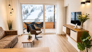 A modern living room with a sofa, a coffee table, a TV, and a balcony with a view of snow-covered mountains. - Alpenstolz #2.05 Comfort (Damüls)