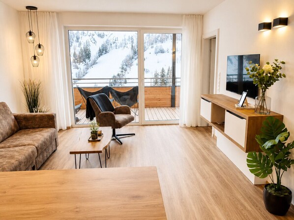 Un salon moderne avec un canapé, une table basse, un téléviseur et un balcon offrant une vue sur des montagnes enneigées.