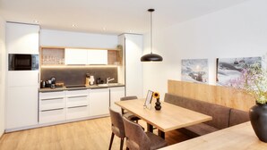 A modern kitchen with white cabinets, a built-in microwave, and a coffee machine. There is a dining area with a wooden table and chairs, and a pendant light above. - Alpenstolz #2.05 Comfort (Damüls)