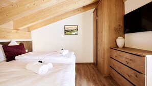 Quarto com teto inclinado, vigas de madeira, cama com roupa branca, cômoda, um vaso com flores e um quadro na parede.
