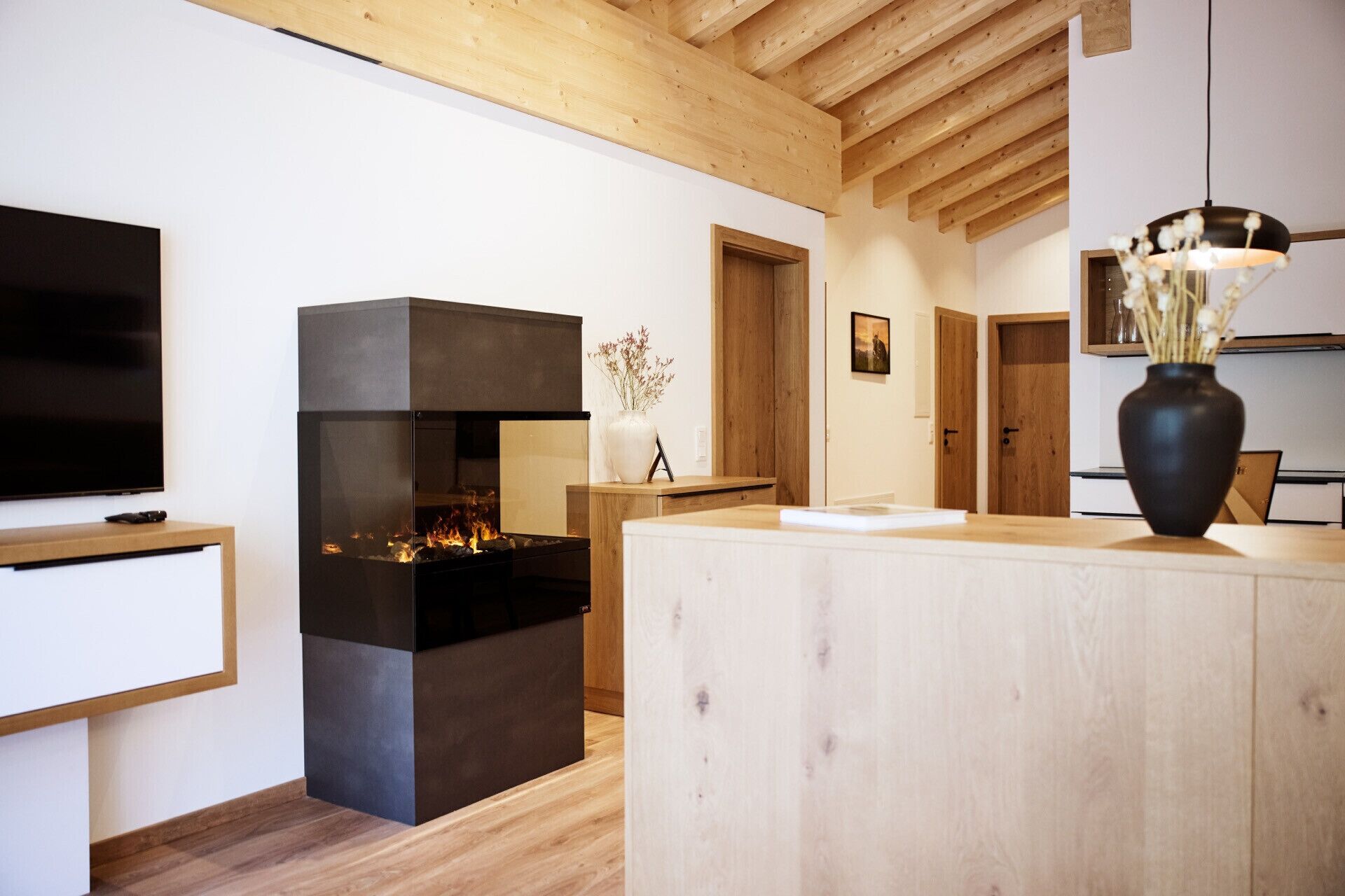 Un salon moderne avec une cheminée, des poutres apparentes en bois au plafond, un téléviseur et un vase rempli de fleurs.