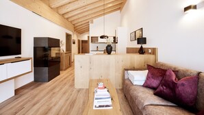 Uma sala de estar moderna com teto de madeira, TV de tela plana, sofá com almofadas, mesa de centro com livros e uma área de cozinha ao fundo.