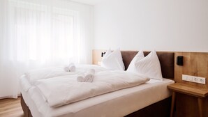 Habitación de hotel con cama, ropa de cama blanca, cabecero de madera, mesita de noche y toma de corriente.