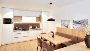 Una cocina moderna con armarios blancos, microondas integrado y cafetera. Cuenta con una zona de comedor con mesa y sillas de madera, y una lámpara colgante.