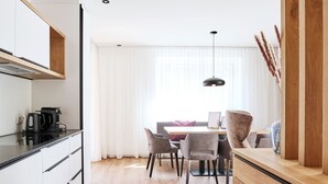 A modern kitchen with white cabinetry and a wooden countertop, a dining area with a table and chairs, and a large window with sheer curtains. - Alpenstolz #3.03 Comfort (Damüls)