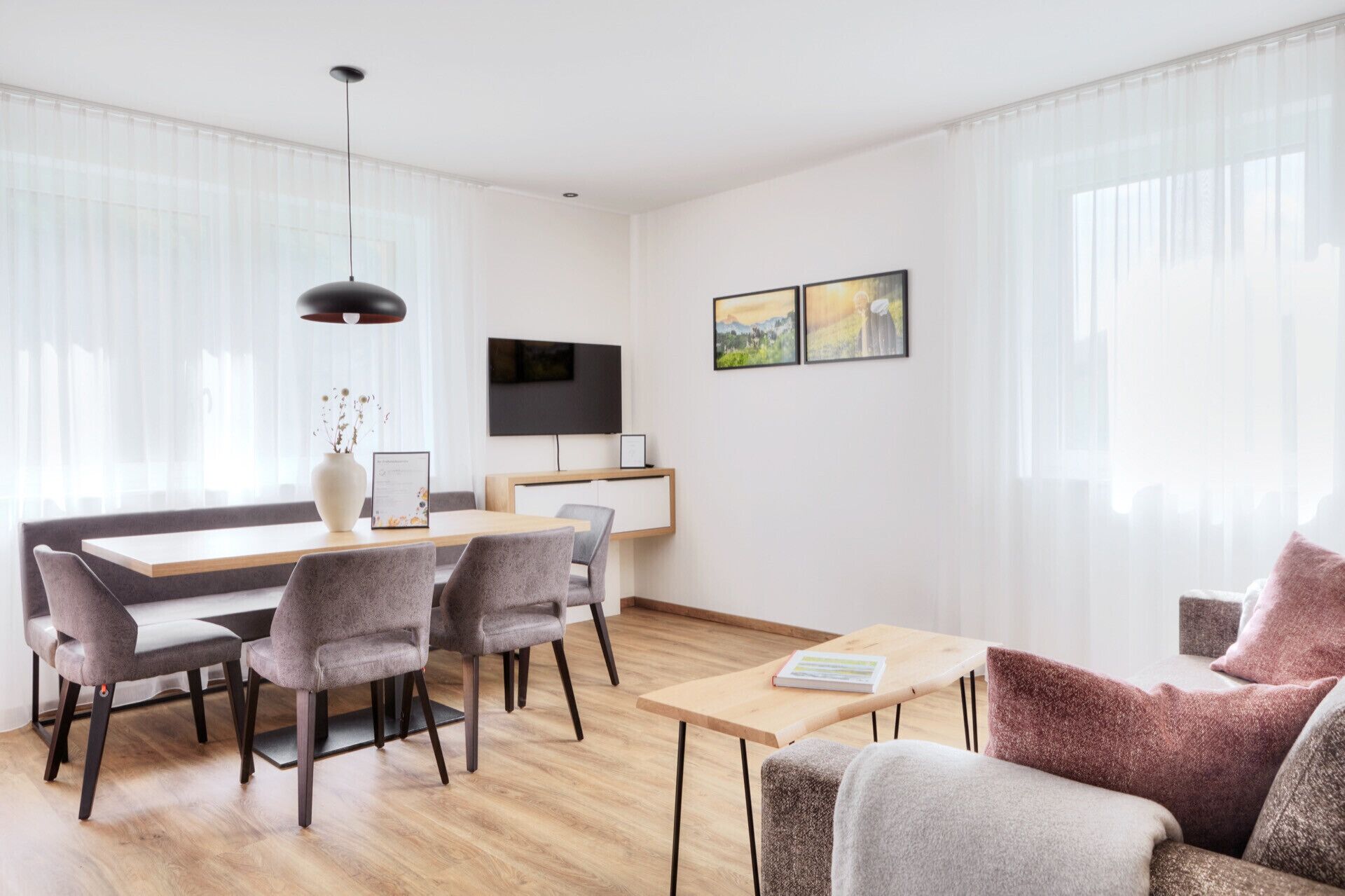 Un soggiorno moderno con zona pranzo, caratterizzato da un tavolo da pranzo in legno, sedie imbottite grigie, un piccolo tavolino e un divano con una coperta rosa.