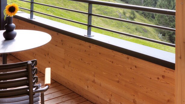 Un balcone con pavimento in legno, un tavolo rotondo con un vaso di girasoli e vista su una catena montuosa.