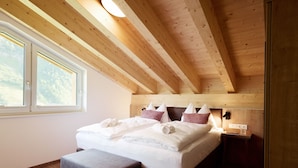 Una camera da letto con soffitto in legno, un letto grande con lenzuola bianche, un comodino e una finestra vista alberi.