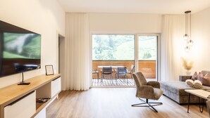 A modern living room with a flat-screen TV, a wooden dining table with chairs, a sofa, and a large window offering a view of a wooden deck.