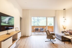 A modern living room with a flat-screen TV, a wooden dining table with chairs, a sofa, and a large window offering a view of a wooden deck. - Alpenstolz #4.01 Comfort (Damüls)