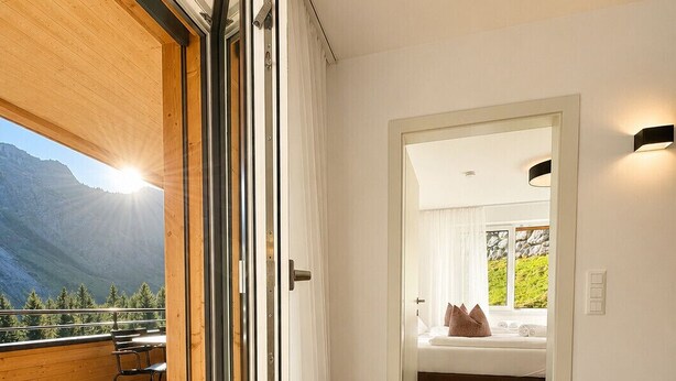 A modern interior with a wooden deck, a bedroom with a bed and curtains, and a view of mountains. - Alpenstolz #4.03 Comfort (Damüls)