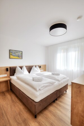A modern hotel room with a large bed, wooden headboard, bedside tables, a ceiling light fixture, a painting on the wall, and a window with sheer curtains.