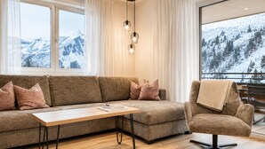 A modern living room with a sofa, a coffee table, and a chair. There are large windows with a view of snow-covered mountains.
