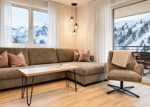 A modern living room with a sofa, a coffee table, and a chair. There are large windows with a view of snow-covered mountains.