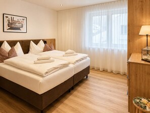 Ein Hotelzimmer mit zwei Betten, einem hölzernen Kopfteil, einem großen Fenster mit durchsichtigen Vorhängen, einem gerahmten Bild an der Wand und einem kleinen Tisch mit einer Lampe.