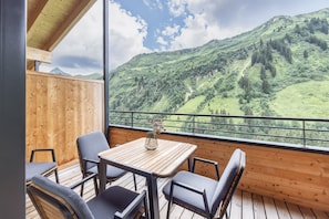 A balcony with a wooden table and chairs, a vase with flowers, and a view of a green valley with mountains.