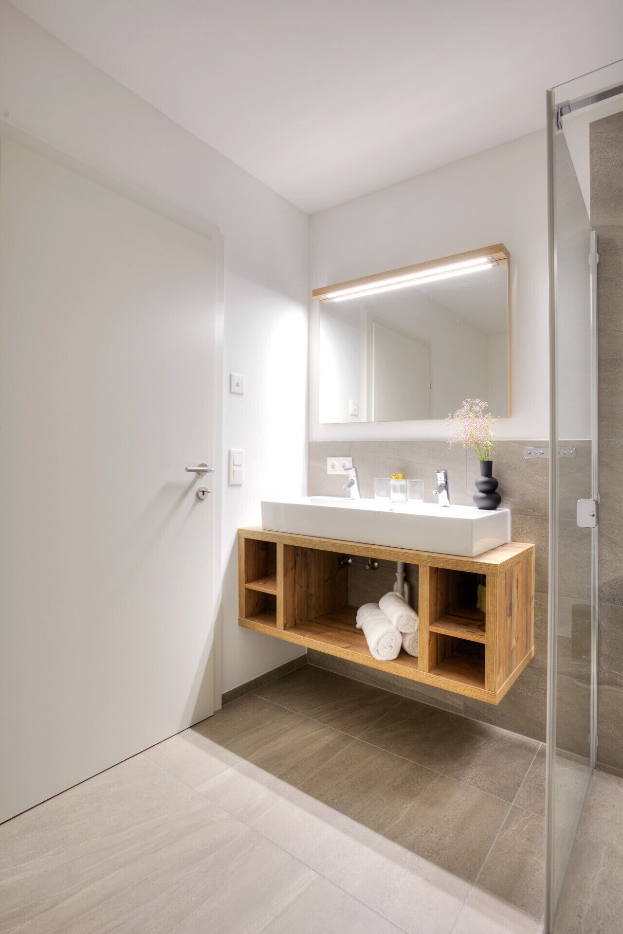Un bany modern amb un tocador de fusta, un mirall gran i un cubicle de dutxa amb portes de vidre.